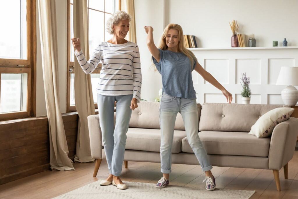 Full length overjoyed middle aged grey haired elderly woman dancing to energetic music with grown up blonde daughter. Happy smiling two generations female family having fun together in living room.