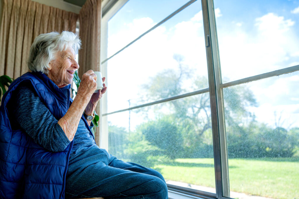 Elderly Senior Caucasian Woman Indoors Holding Coffee or Tea in a Mug and Looking Out the Window Enjoying the View in the Summer